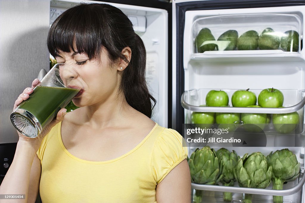 Grimacing mixed race woman drinking healthy drink near refrigerator
