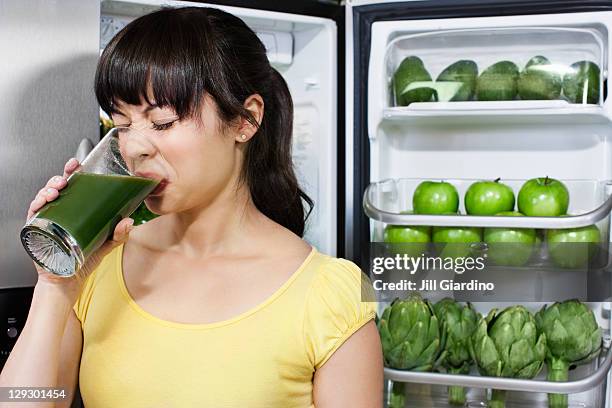 grimacing mixed race woman drinking healthy drink near refrigerator - detox drink stock pictures, royalty-free photos & images