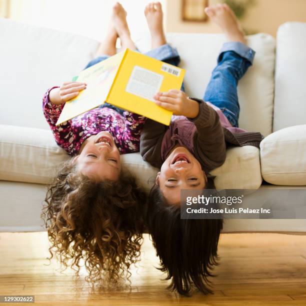 mixed race girls laying upside-down on sofa reading book - kids reading stockfoto's en -beelden