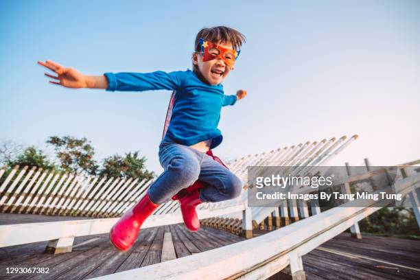 cheerful little girl with a handmade superhero disguise jumping over wooden fence joyfully in park - kindertijd stockfoto's en -beelden