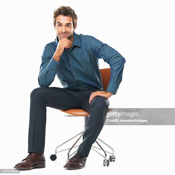 studio shot of young business man sitting on chair with hand on chin and smiling - kin in de hand stockfoto's en -beelden