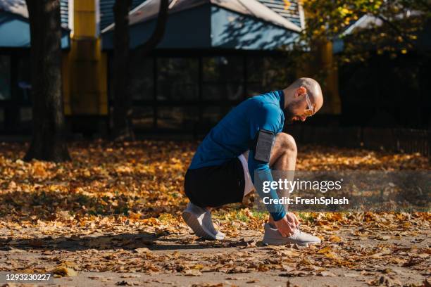 handsome young man in a public park, making a short break from exercising to tie his shoelace - blue november stock-fotos und bilder