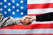 cropped view of soldier shaking hand with civilian man near american flag on blurred background