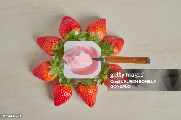 above view of a spoon with red yoghurt on a yoghurt cup made of plastic with eitght strawberries with green leaves around the cup on a white backgrounds. horizontal photo. - joghurtbecher stock-fotos und bilder