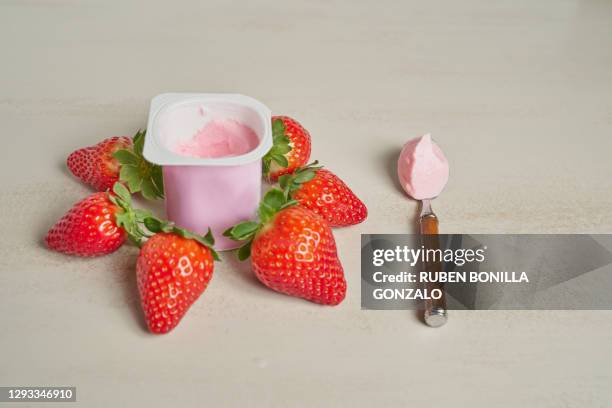 front view of a spoon with red yoghurt on a yoghurt cup made of plastic with various strawberries with green leaves around the cup on a white backgrounds. horizontal photo. - joghurtbecher stock-fotos und bilder