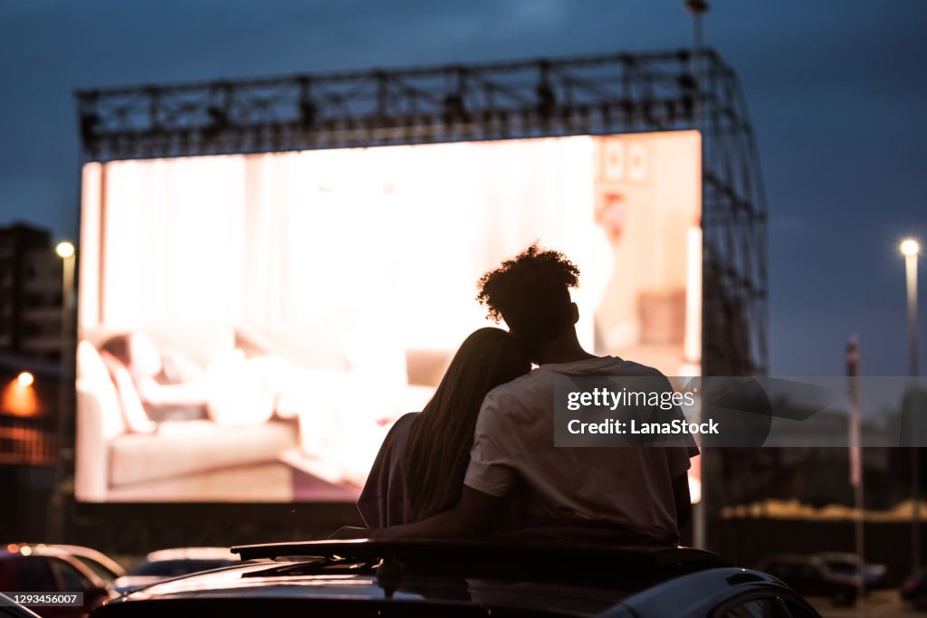 Silhouetted view of attractive young couple, boy and girl embracing, spending time together, sitting in the car while watching a movie in a drive in cinema