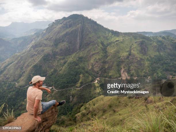 young man sitting on rock on top of beautiful mountain range in sri lanka admiring the green lush landscape - adams peak stock pictures, royalty-free photos & images