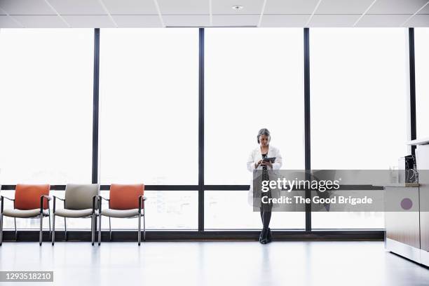 female doctor using digital tablet while standing against window in hospital - hospital waiting room stock pictures, royalty-free photos & images