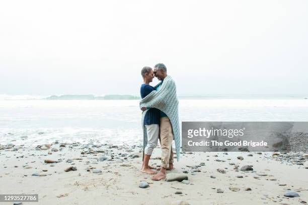 happy mature woman embracing male partner at beach against clear sky - happy couple cuddle stockfoto's en -beelden
