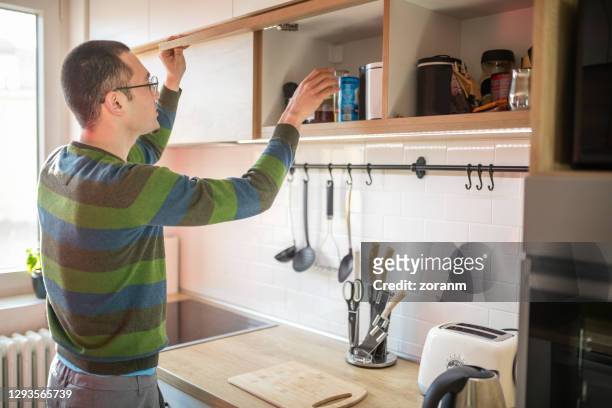 young man opening upper cabinet and fetching jar - jars kitchen stock pictures, royalty-free photos & images