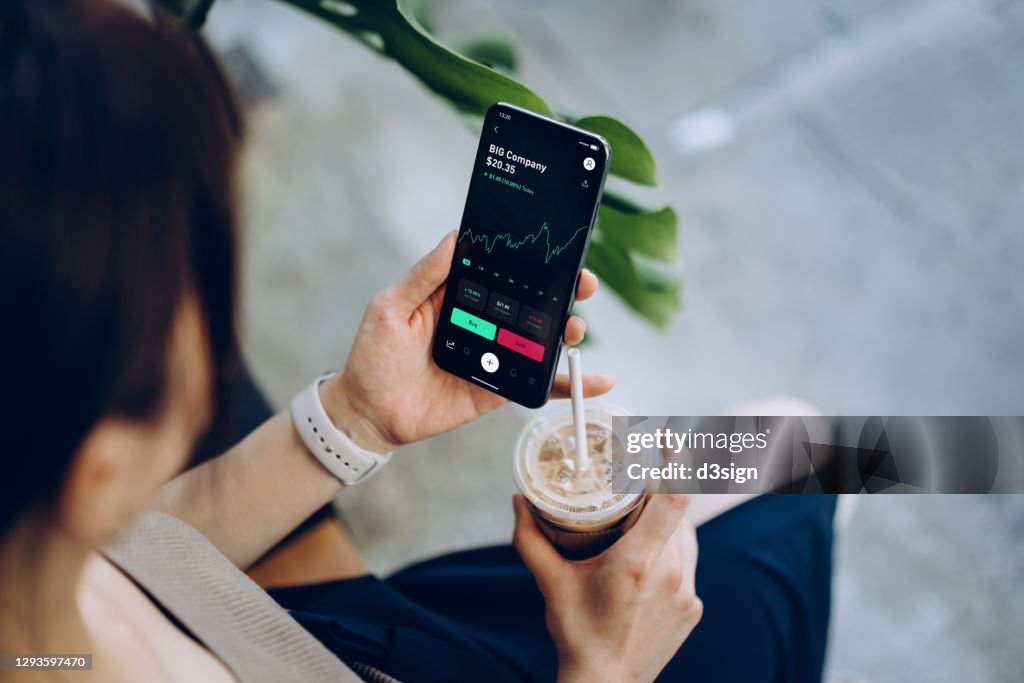 Over the shoulder view of young Asian woman checking financial trading data with mobile app on smartphone while sitting outdoors in a cafe, drinking coffee. Business on the go