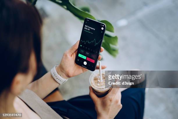 over the shoulder view of young asian woman checking financial trading data with mobile app on smartphone while sitting outdoors in a cafe, drinking coffee. business on the go - börsenhausse stock-fotos und bilder