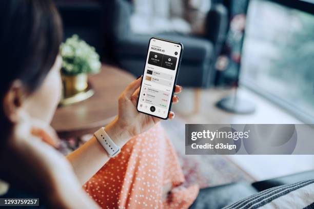 over the shoulder view of young asian woman relaxing at home, sitting on the sofa in the living room, managing online banking with mobile app on smartphone. transferring money, paying bills, checking balance. technology makes life so much easier - electronic banking fotografías e imágenes de stock