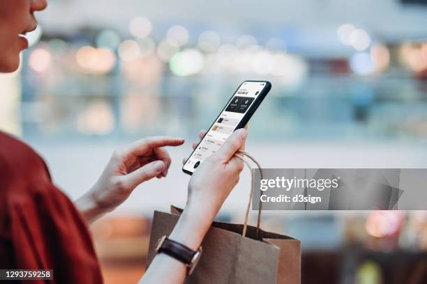 cropped shot of young asian woman holding a paper shopping bag, managing online banking with mobile app on smartphone. transferring money, paying bills, checking balance while shopping in a shopping mall. technology makes life so much easier - concept store fotografías e imágenes de stock