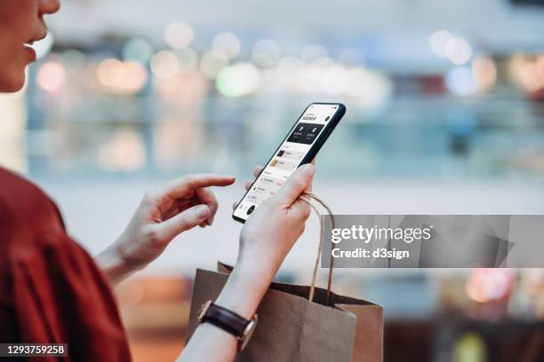 cropped shot of young asian woman holding a paper shopping bag, managing online banking with mobile app on smartphone. transferring money, paying bills, checking balance while shopping in a shopping mall. technology makes life so much easier - e commerce asian stock pictures, royalty-free photos & images