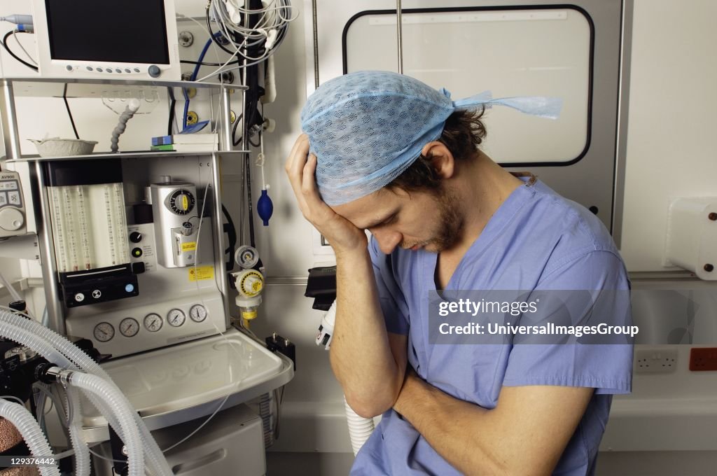 Male doctor, wearing green surgical cap and gown, showing signs of stress and tiredness