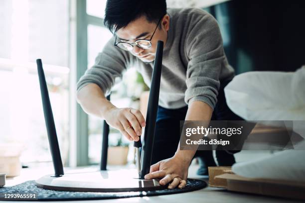young asian man assembling furniture with a screwdriver, he is setting up a wooden coffee table in newly refurbished apartment against sunlight - furniture maker foto e immagini stock