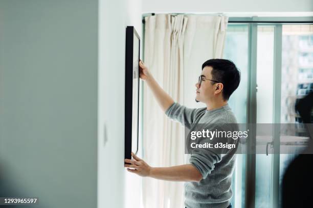 young asian man holding a picture frame and hanging the painting on the wall decorating in his newly refurbished apartment - hangen stock-fotos und bilder