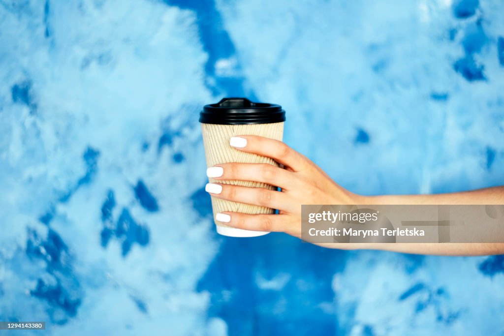 Paper cup in female hands against a blue wall.