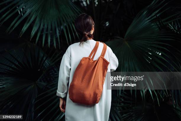 rear view of young asian woman relaxing in a park. she is carrying a brown reusable bag against green nature plants. responsible shopping, zero waste and sustainable lifestyle concept - mode stock-fotos und bilder