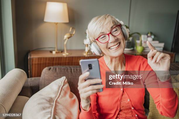la femme aînée gaie utilise le téléphone portable et le casque à la maison - une seule femme senior photos et images de collection