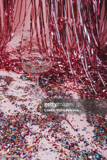 a festive glass with champagne and sequins stands on a pink background of tinsel and confetti - lametta stock-fotos und bilder