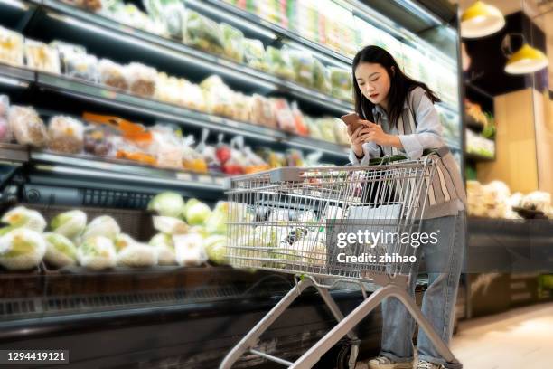 young asian female customer pushing shopping cart with mobile phone in supermarket - shopping trolleys stockfoto's en -beelden