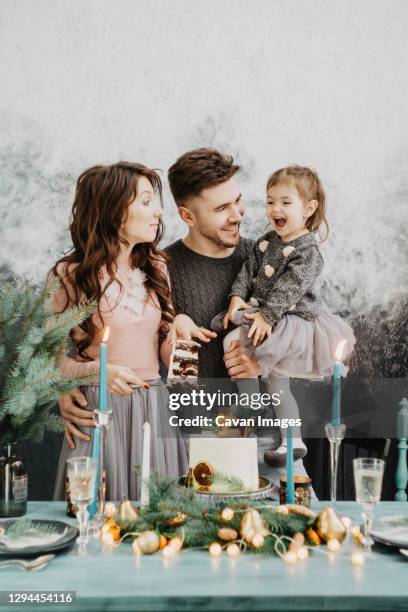 young family with a daughter in festive outfits at a served table with candles, garlands, sparklers and a cake near the christmas tree on new years eve - christmas family tree stock-fotos und bilder