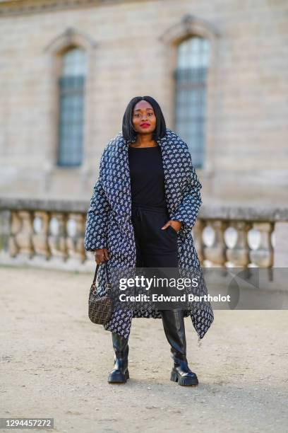 Carrole Sagba wears a long winter puffer coat with printed monograms from Saks Potts, a black pullover from Acne Studios, a brown monogram print...