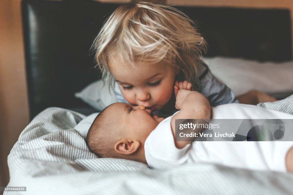 Little brother hugging his newborn sister. Toddler kid meeting new sibling. Cute boy and new born baby girl relax in a white bedroom. Family with children at home. Love, trust and tenderness