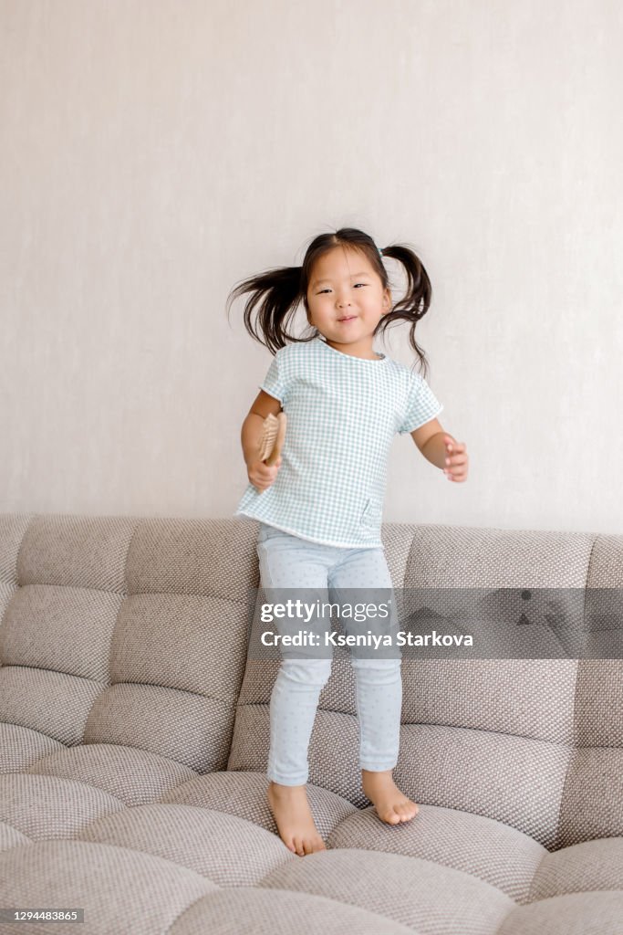 Little asian girl jumping on a light sofa in the living room dancing with a comb in her hands