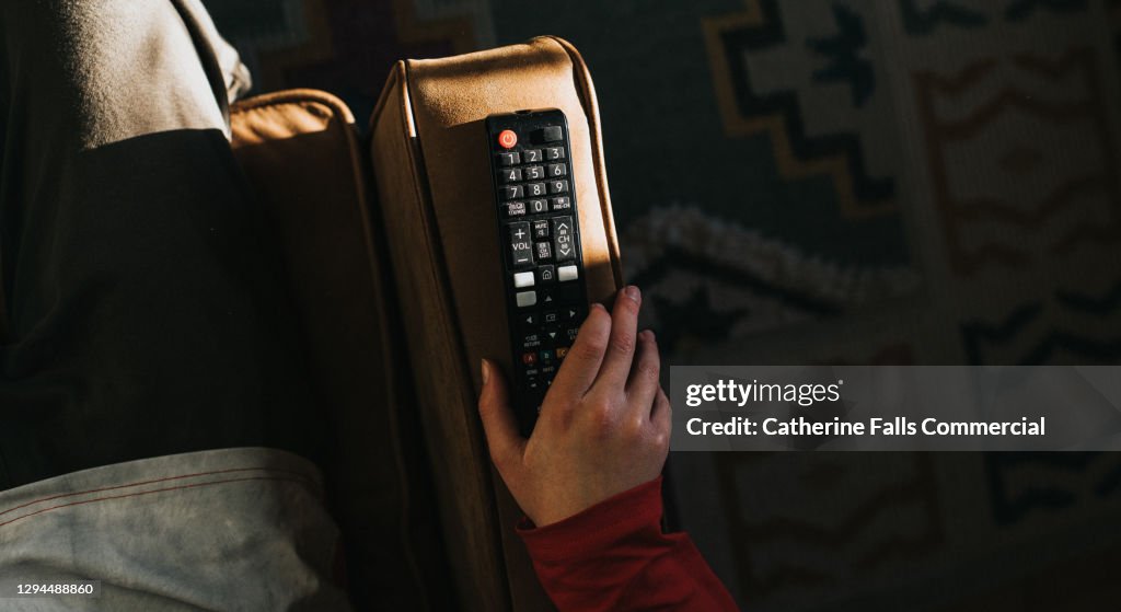 A hand holding a Television Remote Control on the Arm of a Sofa