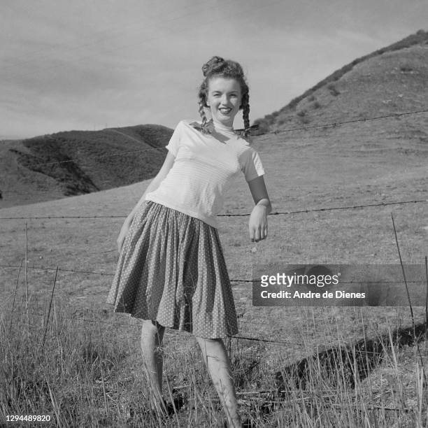 Portrait of American actress and model Marilyn Monroe with pigtails as dressed in a short-sleeve top and a skirt, as she poses in a field, Northern...