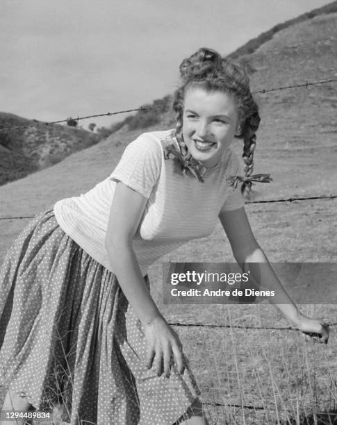 Portrait of American actress and model Marilyn Monroe with pigtails as dressed in a short-sleeve top and a skirt, as she poses in a field, Northern...