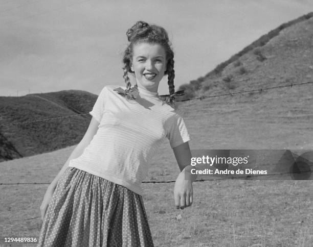 Portrait of American actress and model Marilyn Monroe with pigtails as dressed in a short-sleeve top and a skirt, as she poses in a field, Northern...