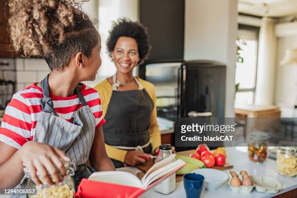 daughter helps her mother in the kitchen and reads a recipe from a recipe book - cookbook stock pictures, royalty-free photos & images