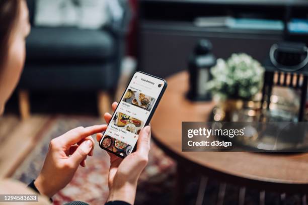 close up of young asian woman using meal delivery service and ordering food online with mobile app on smartphone while sitting on the sofa in the living room at a cozy home - women reservation stock pictures, royalty-free photos & images