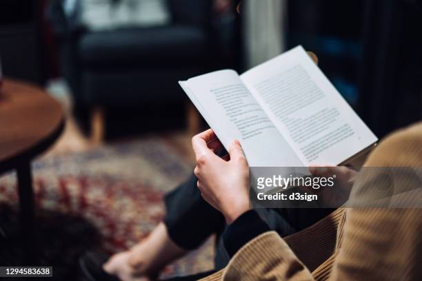 cropped shot of young asian woman taking a break from technology, relaxing and reading book on the sofa at a cozy home in the evening - boek stockfoto's en -beelden