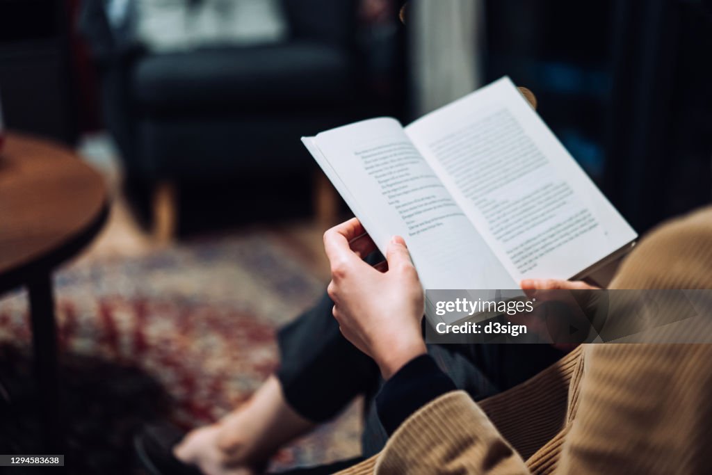 Cropped shot of young Asian woman taking a break from technology, relaxing and reading book on the sofa at a cozy home in the evening