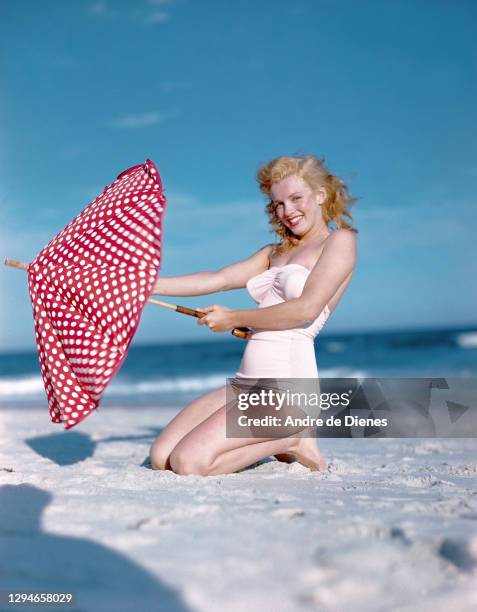 Portrait of American actress and model Marilyn Monroe , in a white swimsuit, as she poses with a red, polka dot umbrella on Long Island's Tobay...