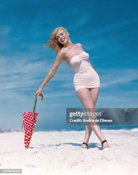Portrait of American actress and model Marilyn Monroe , in a white swimsuit, as she poses with a red, polka dot umbrella on Long Island's Tobay...