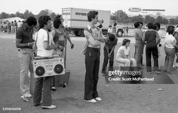 View of a group of people waiting in line, one with a large 'boom box' radio slung around his shoulder, in Flushing Meadows Park, in the Corona...