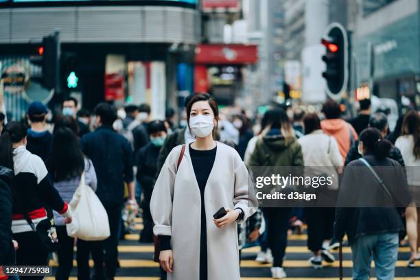 portrait of young asian woman with protective face mask standing in the middle of busy downtown city street amidst crowd of pedestrians passing by, looking at the camera, with highrise city buildings and busy traffic in background - alone in a crowd sad stockfoto's en -beelden