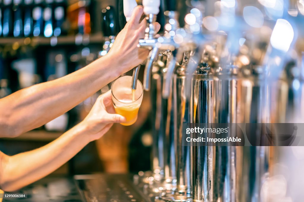 Male hands poured beer from the tap. Pouring beer for client. Side view of young bartender pouring beer while standing at the bar counter.
