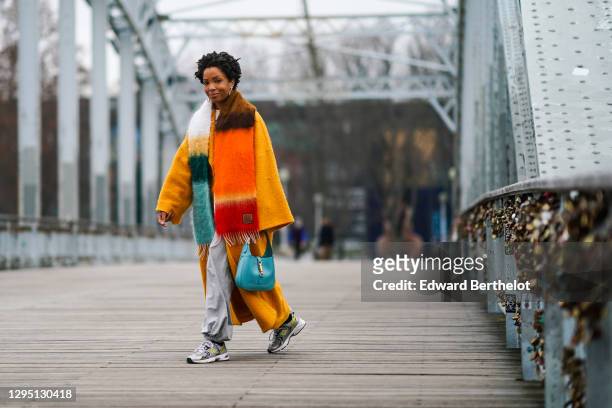 Ellie Delphine wears golden earrings, a multi colored orange and blue striped wool Loewe fringed scarf, an orange wool oversized long coat with...