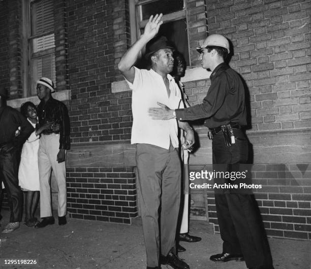 An African American man raises his arm as he is given a body search by a police officer, as a young African American woman watches in tears, on the...