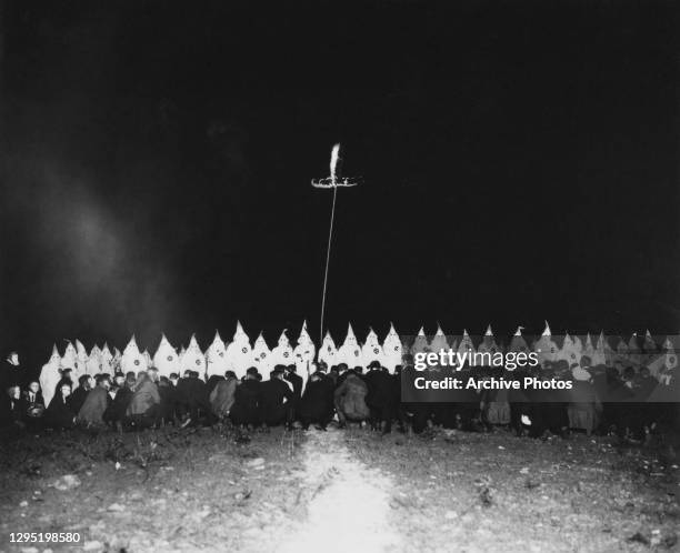 Hooded Klansmen stand in a line with a burning cross behind them, and a group of uncostumed people sitting on the ground before them during a Ku Klux...