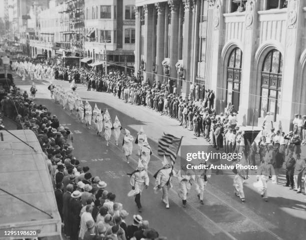 Flag bearer, carrying the Stars-and-Stripes, as they lead a Ku Klux Klan procession through a city centre, watched by crowds on either side of the...