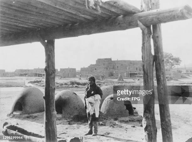 Native American Hopi woman standing before the clay and mud ovens in which she bakes bread, with adobe dwellings in the background, on the Hopi...