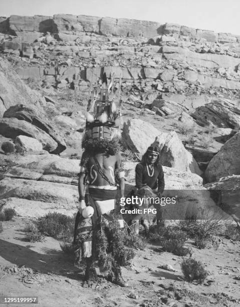 Native American Hopi man stands in ceremonial costume, with a second tribesperson sits behind with a dog, against the rugged landscape of the Hopi...